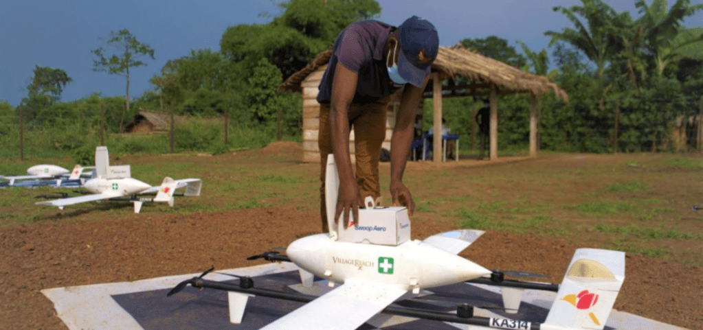 a photos of a yard in Africa with three drones sitting on the ground. A man is loading supplies into one drone. 